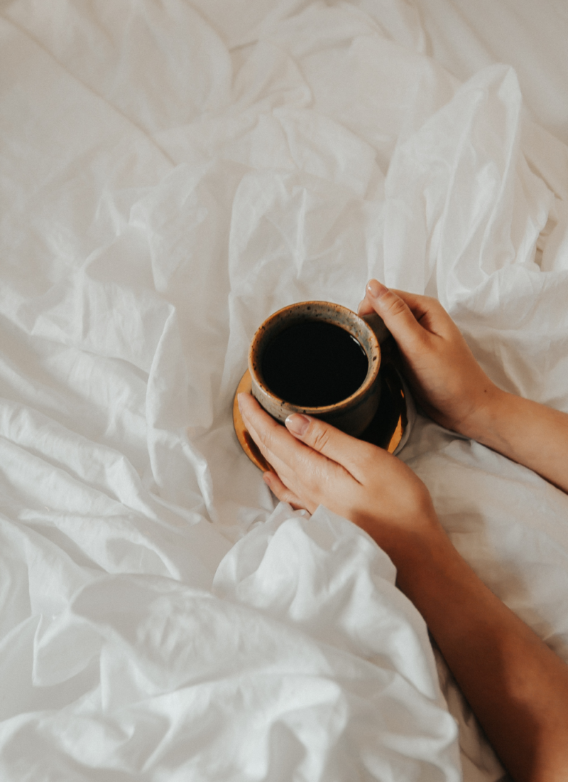 Woman holding a cup of coffee on top of her bed to wake up and be productive in the morning