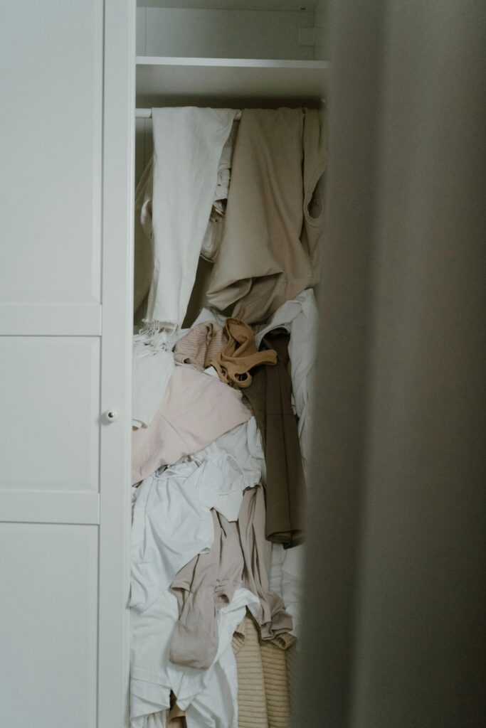 The picture shows a closet with unorganized white, brown and beige color clothes.